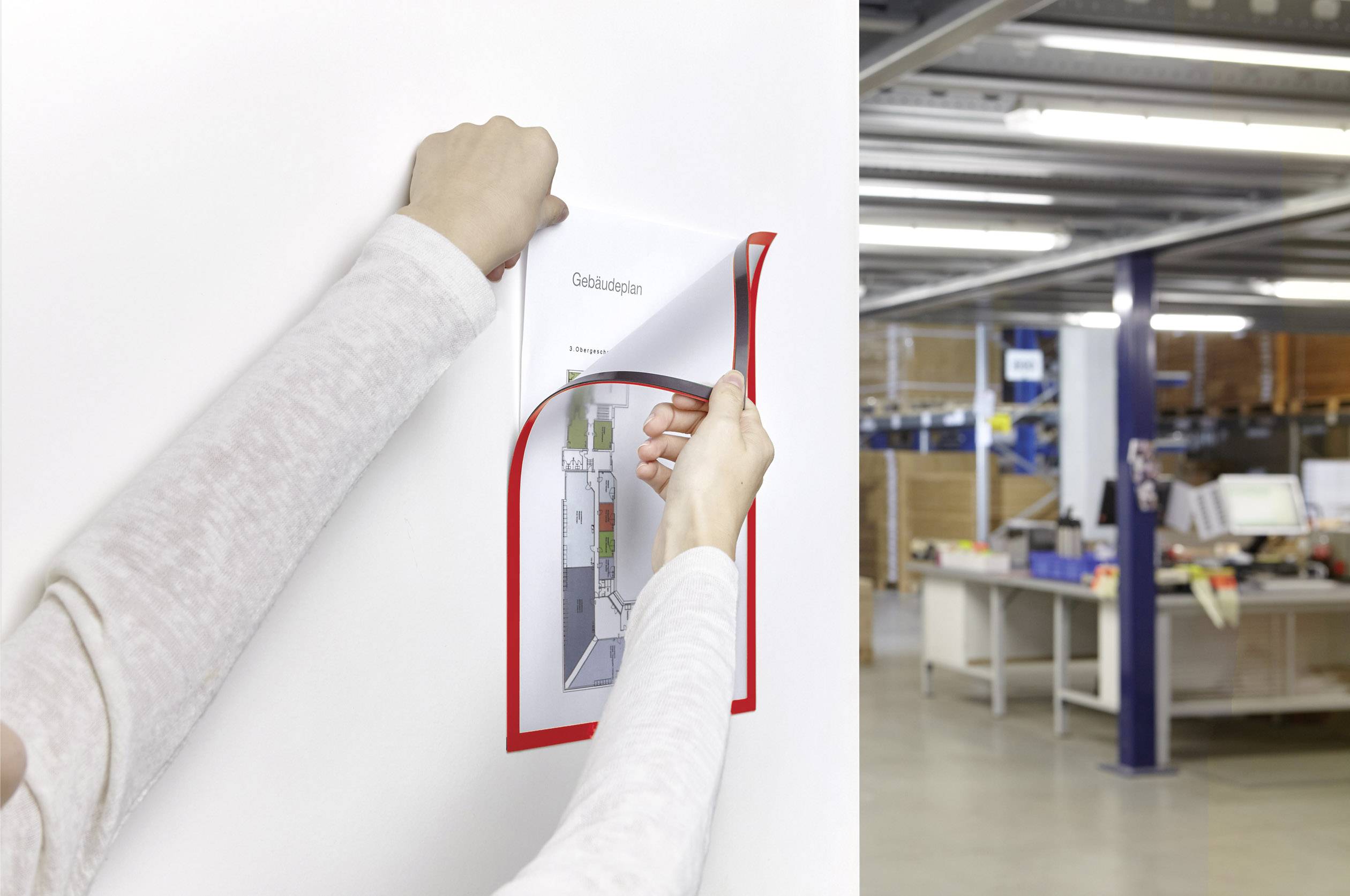 Une personne attache un document dans un cadre rouge sur un mur blanc. À l'arrière-plan, on aperçoit un bureau avec des bureaux.