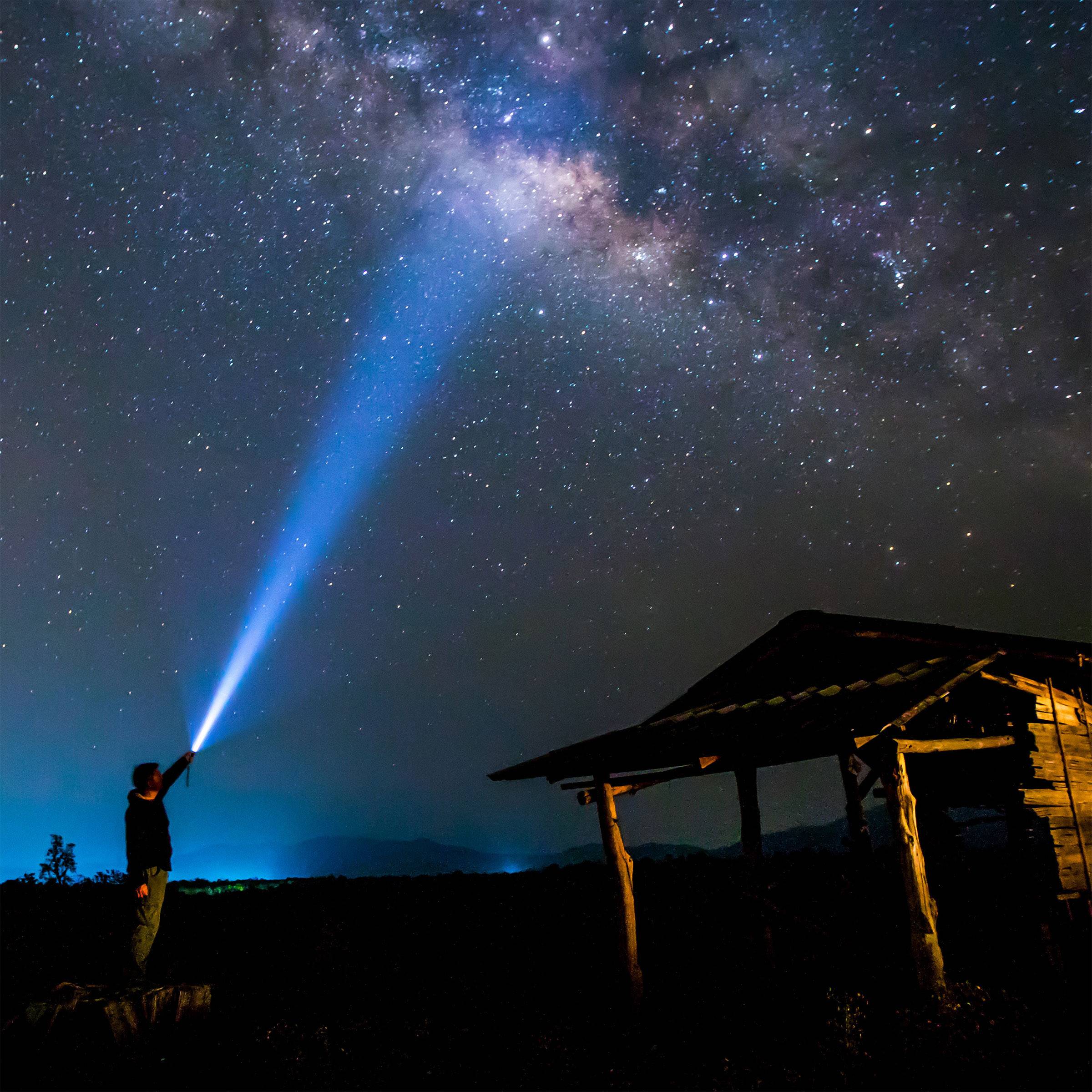 Une personne se tient dans l'obscurité et éclaire le ciel étoilé clair avec une lampe de poche, à côté d'une structure en bois.