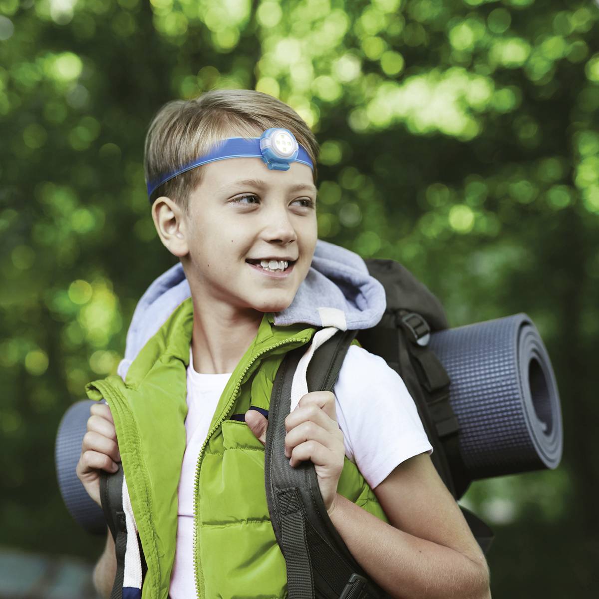 Un garçon à l'extérieur porte un bandeau frontal et un sac à dos avec un matelas de camping. Il sourit, entouré de nature verdoyante.