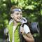 Un garçon à l'extérieur porte un bandeau frontal et un sac à dos avec un matelas de camping. Il sourit, entouré de nature verdoyante.