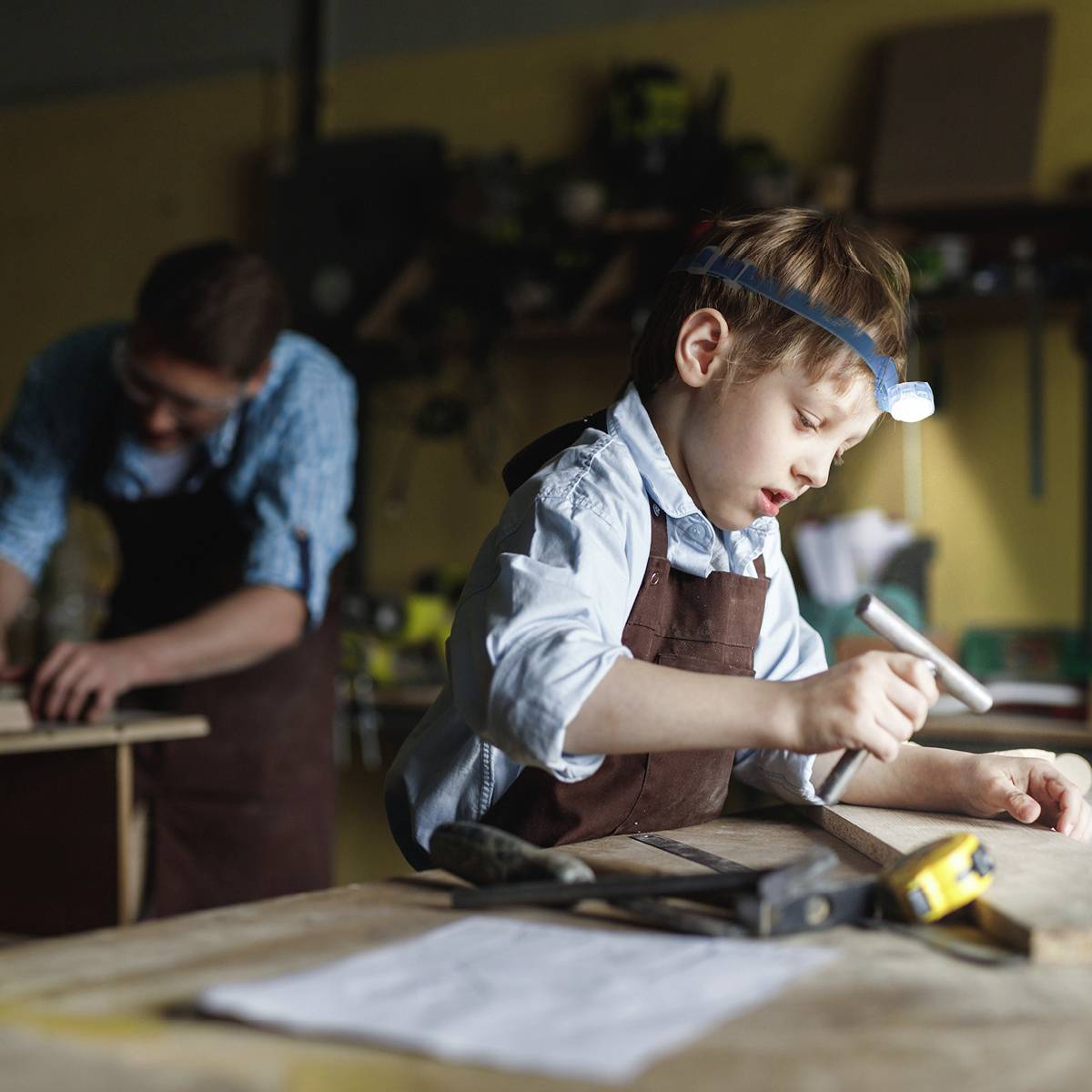 Un enfant travaille avec concentration sur un morceau de bois, tenant un stylo pour mesurer. À l'arrière-plan, une autre personne travaille sur un projet similaire.