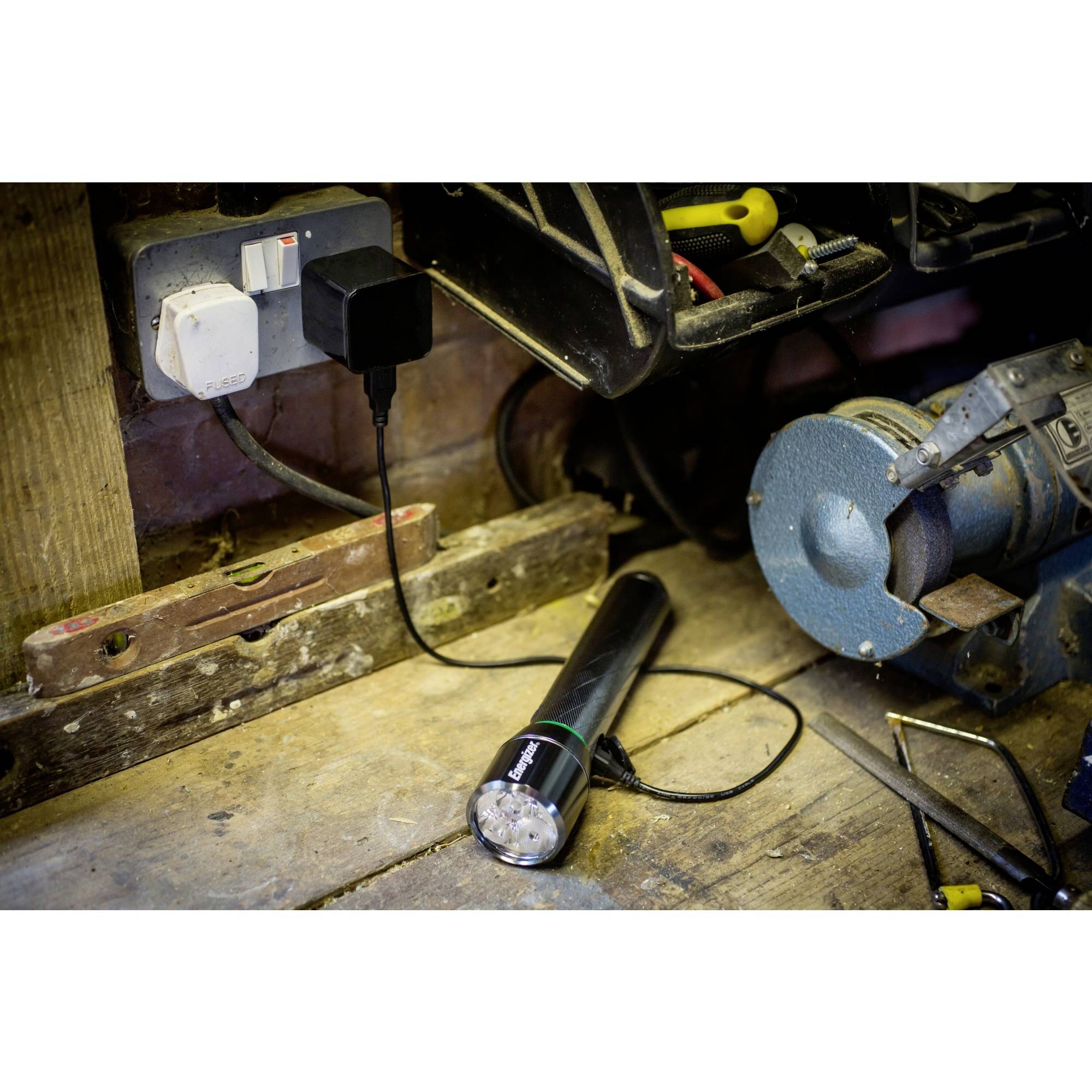 Une lampe de poche est en train de se recharger dans un atelier. Elle est posée sur un établi à côté d'un chargeur, d'outils et d'une meuleuse.