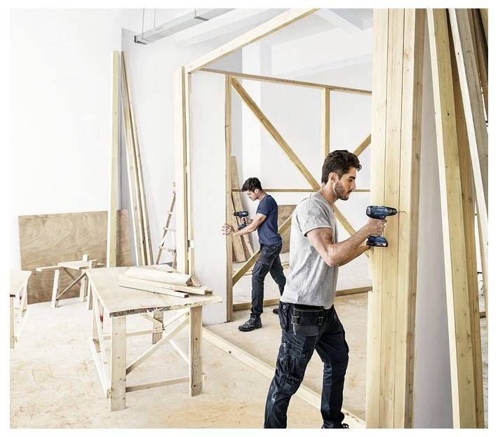 Deux personnes utilisent des perceuses pour assembler une structure en bois dans un atelier, entourées de diverses pièces de bois et d'outils.