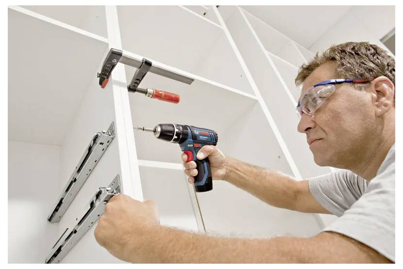 Une personne installe des glissières de tiroir métalliques sur des étagères en bois à l'aide d'une perceuse électrique, en portant des lunettes de protection pour la sécurité.