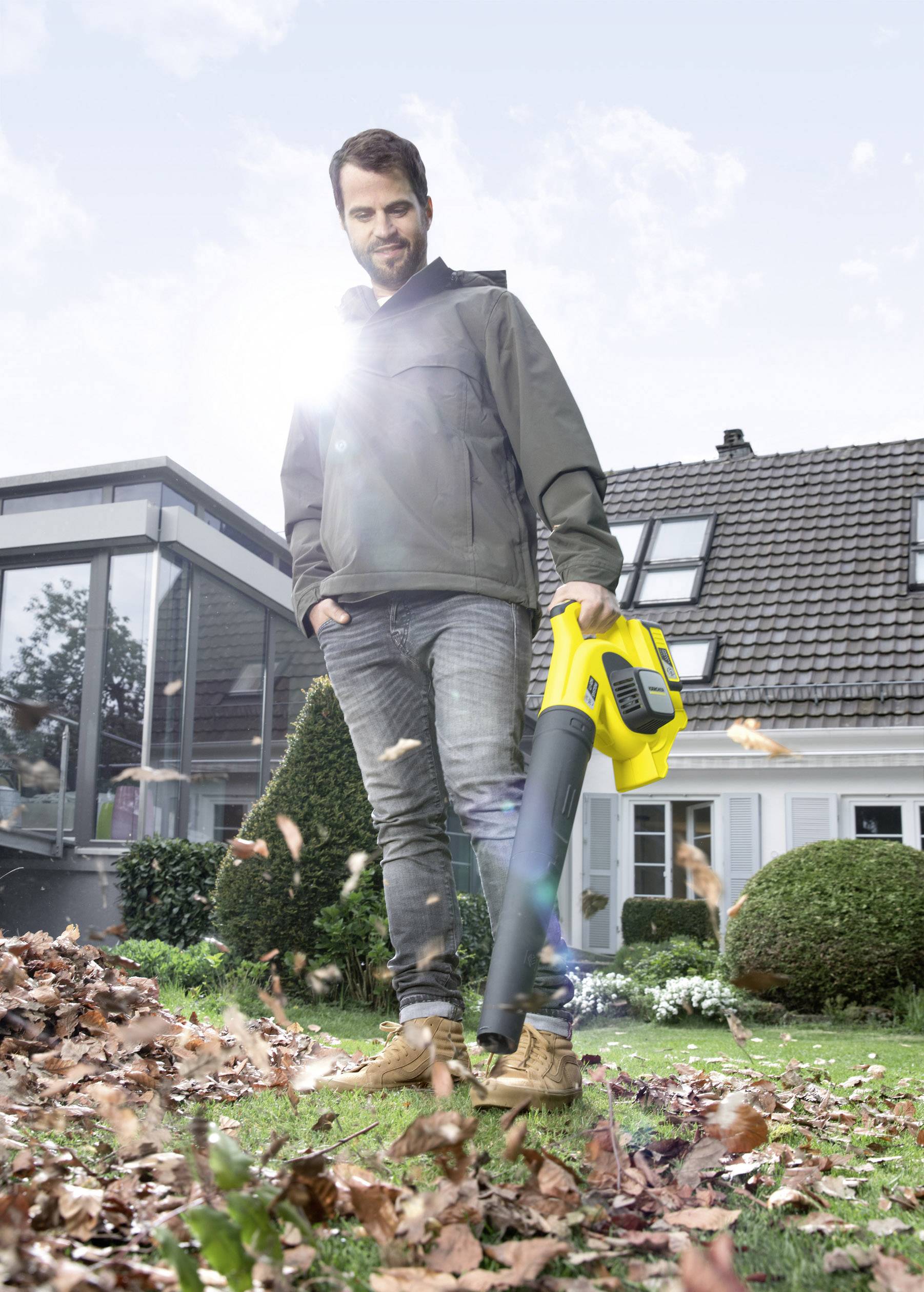 Un homme se tient dans le jardin et utilise un souffleur de feuilles jaune pour enlever les feuilles d'automne de la pelouse devant une maison.