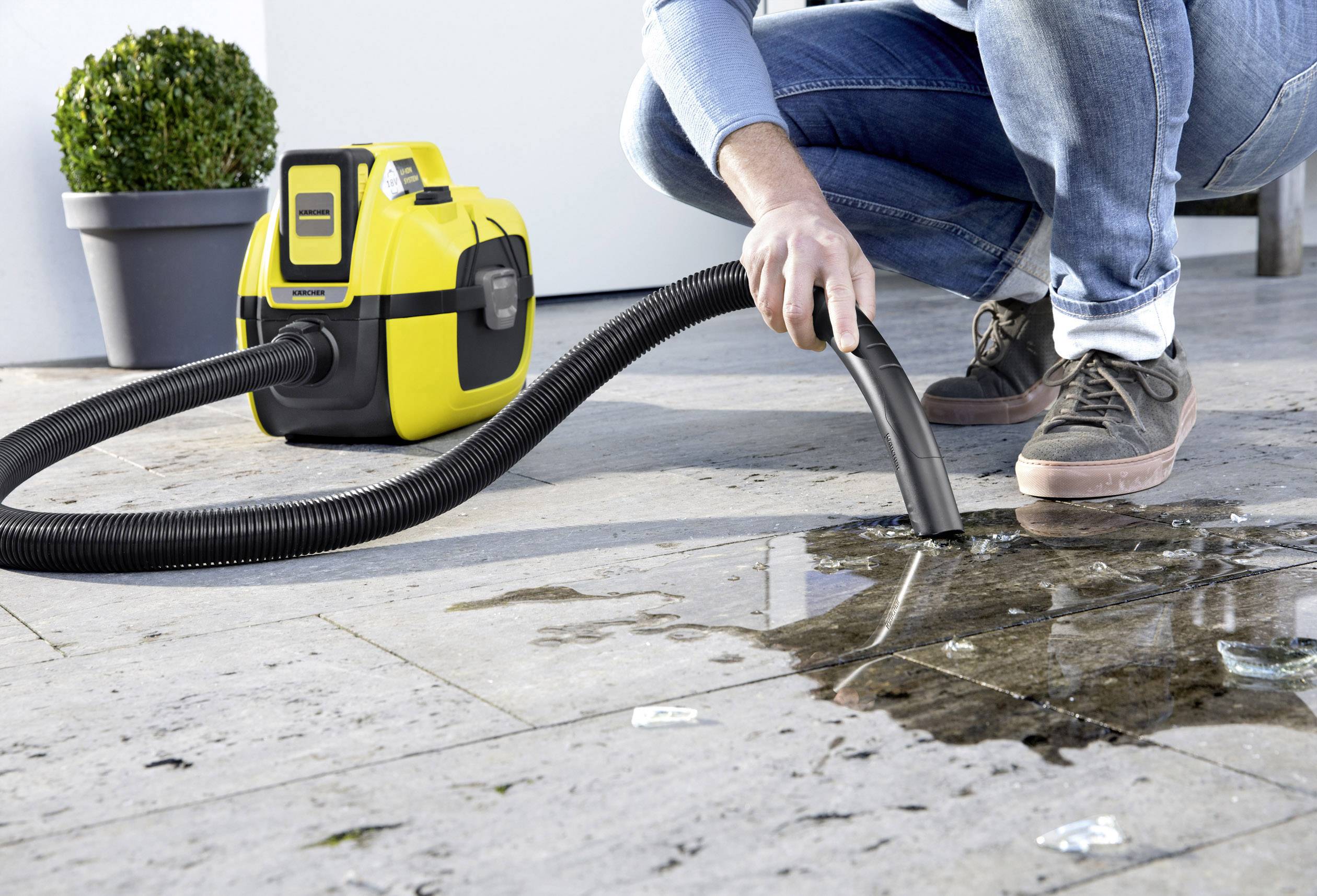 Une personne aspire de l'eau sur un sol carrelé à l'aide d'un aspirateur eau et poussière jaune. À l'arrière-plan, une plante est placée dans un pot.
