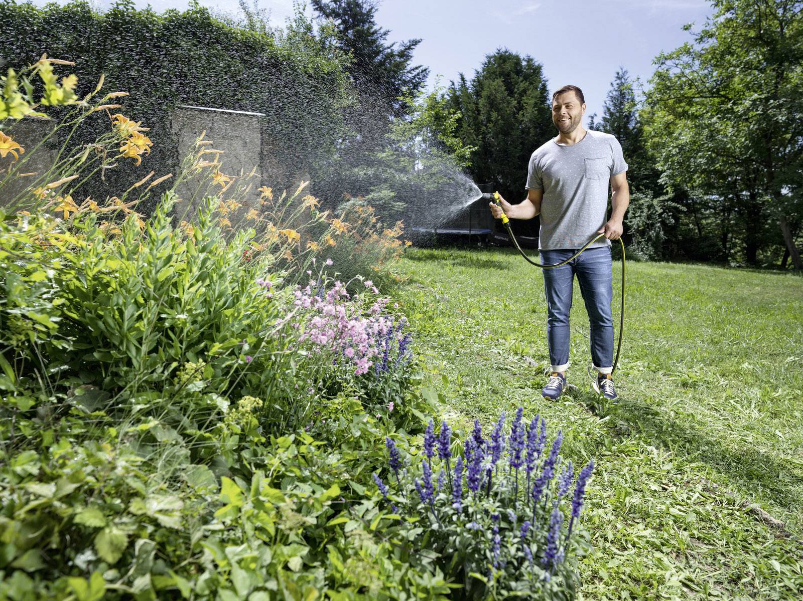 Un homme arrose un massif de fleurs avec un tuyau d'arrosage dans un jardin verdoyant par temps ensoleillé.