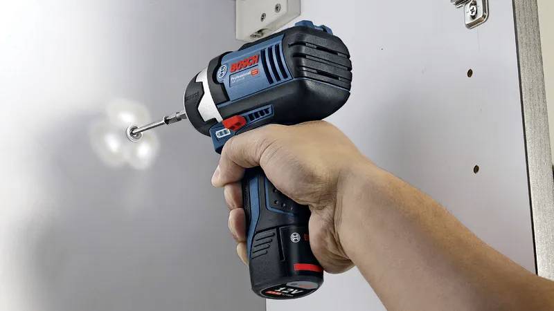 Une main utilisant une perceuse sans fil pour visser une vis dans une surface blanche, démontrant l'utilisation d'un outil et son application pratique.