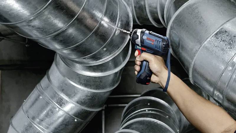 Une personne utilisant une perceuse sans fil bleue pour fixer un conduit métallique dans un environnement industriel, illustrant l'application de l'outil dans le domaine de la construction.