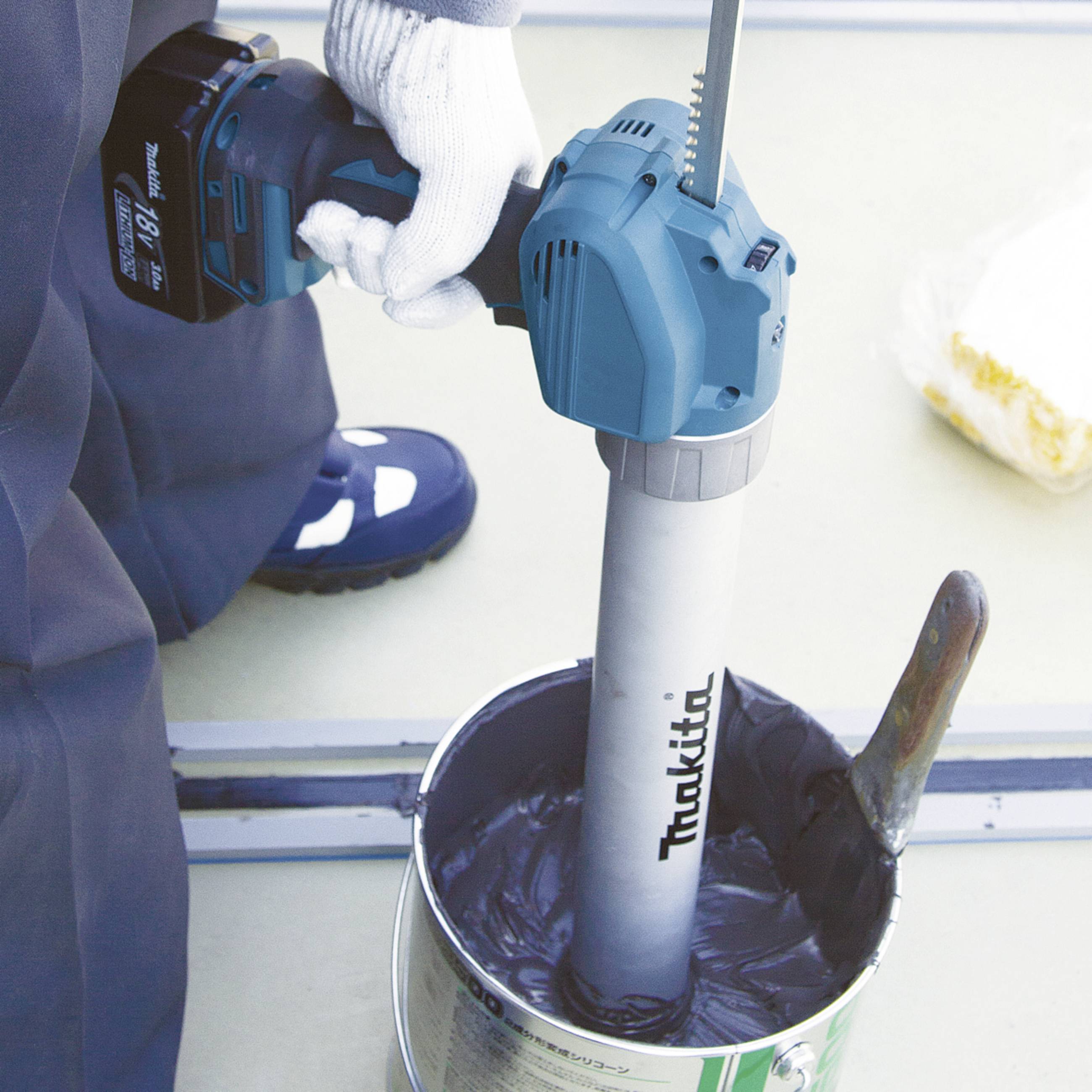 'Un ouvrier utilise une scie sauteuse sans fil, dont la lame est plantée dans un seau de peinture. Des vêtements de travail et des gants de protection sont visibles.'