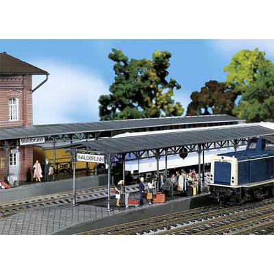 Scène de train miniature dans une gare avec un train et des personnages. Des personnes se tiennent sur le quai avec leurs bagages devant un train portant l'inscription 'WaldbRUNN'.