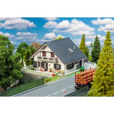 Une maquette de maison se trouve dans un paysage avec des arbres en arrière-plan. Un camion-miniature chargé de grumes roule sur la route.