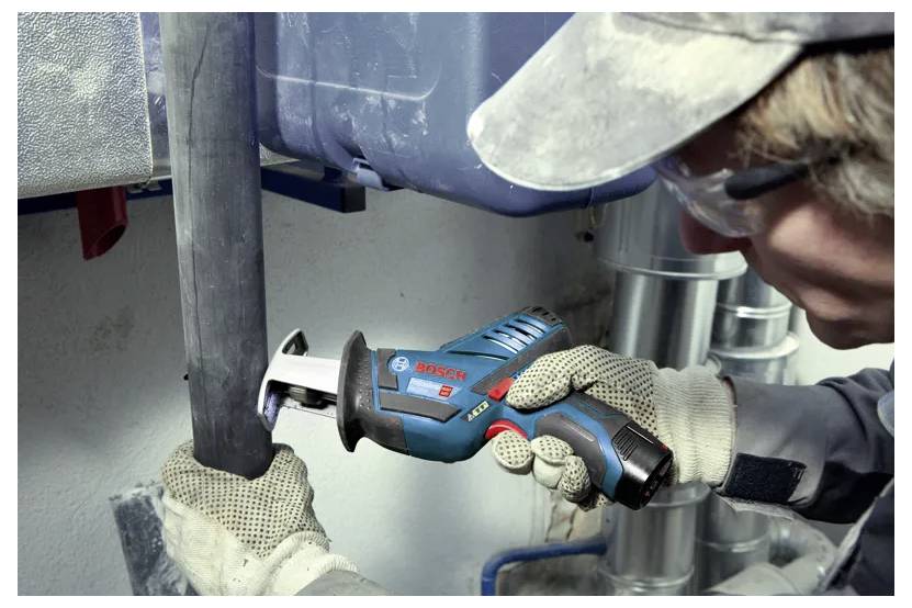 Un travailleur utilise un outil électrique Bosch pour couper un tuyau, illustrant sa concentration sur un travail de construction. L'ouvrier porte des gants de protection et des lunettes de sécurité.