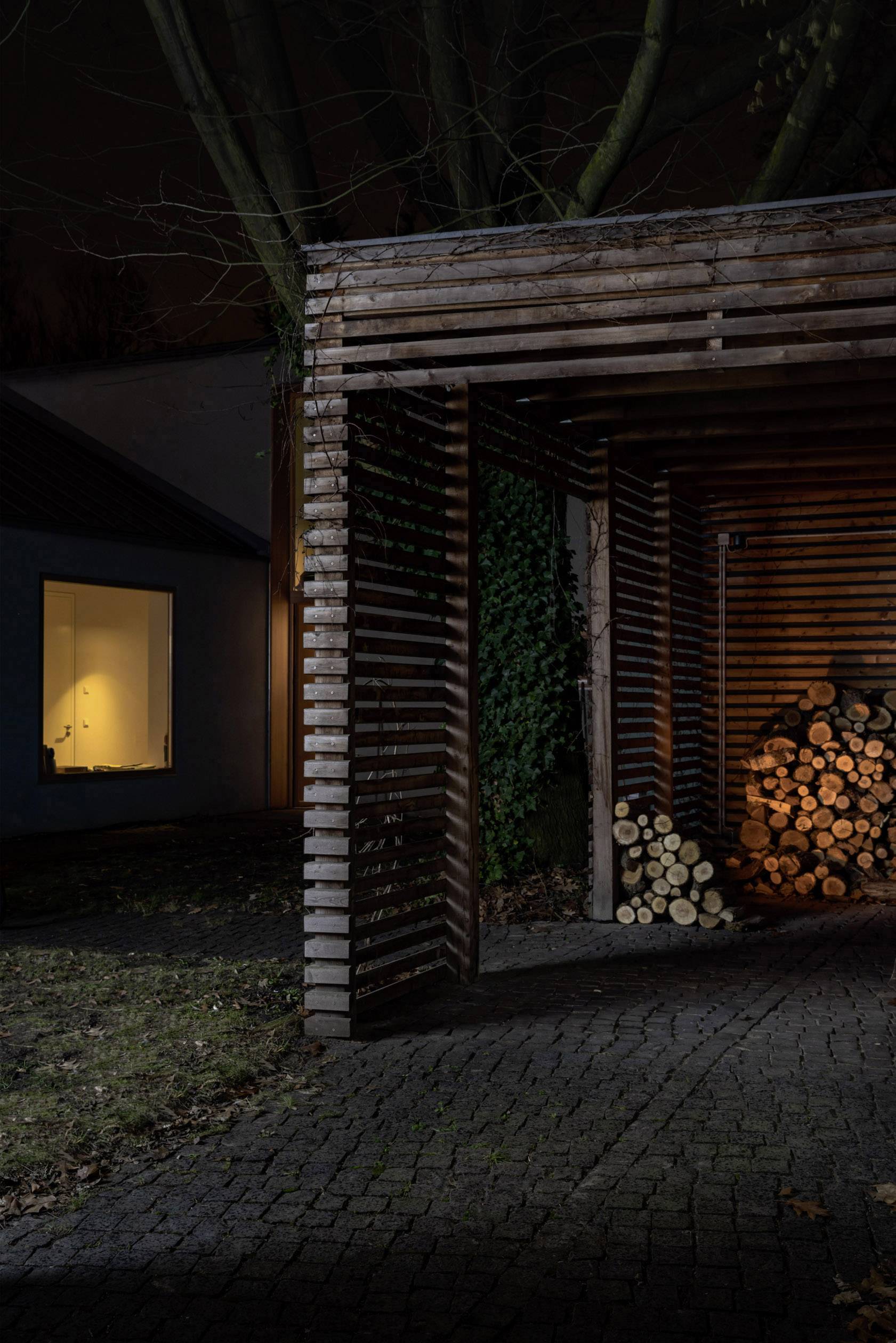 Abri à bois la nuit, avec du bois de chauffage empilé. Une fenêtre éclairée d'une maison en arrière-plan.