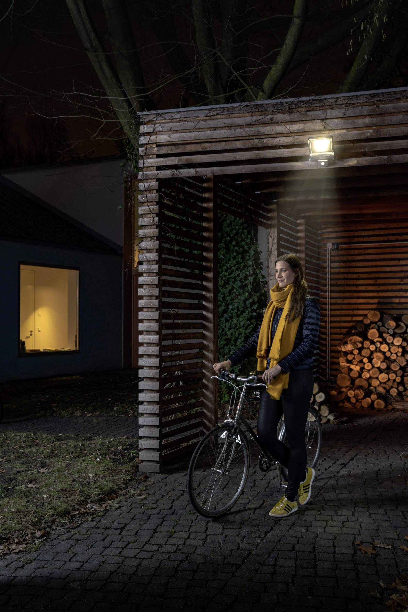 Femme portant une écharpe jaune et des chaussures jaunes, debout avec son vélo dans une allée en bois éclairée. À l'arrière-plan, des bûches sont empilées.