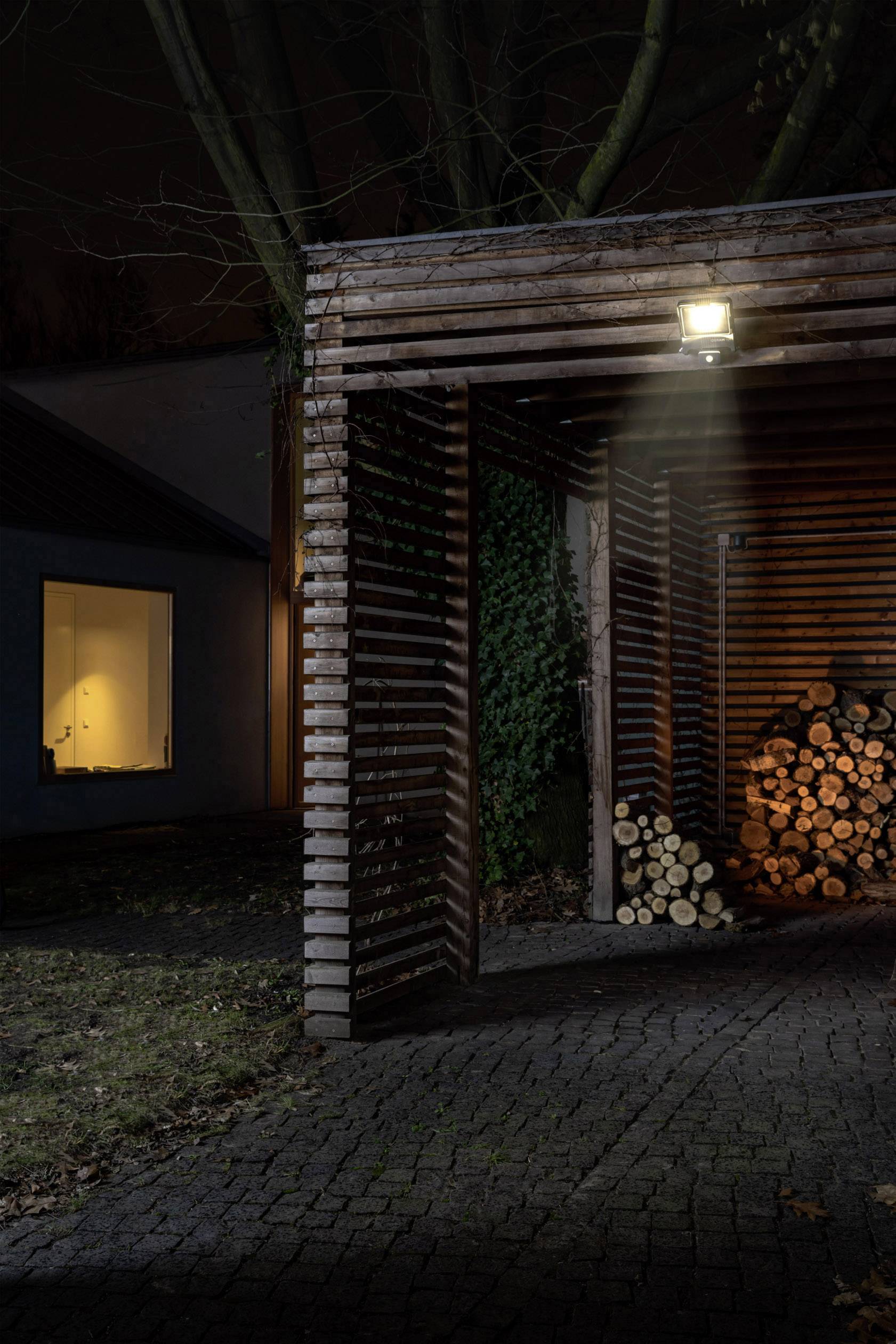 Un abri en bois éclairé avec du bois de chauffage empilé. À l'arrière-plan, une fenêtre illuminée d'une maison dans la nuit.