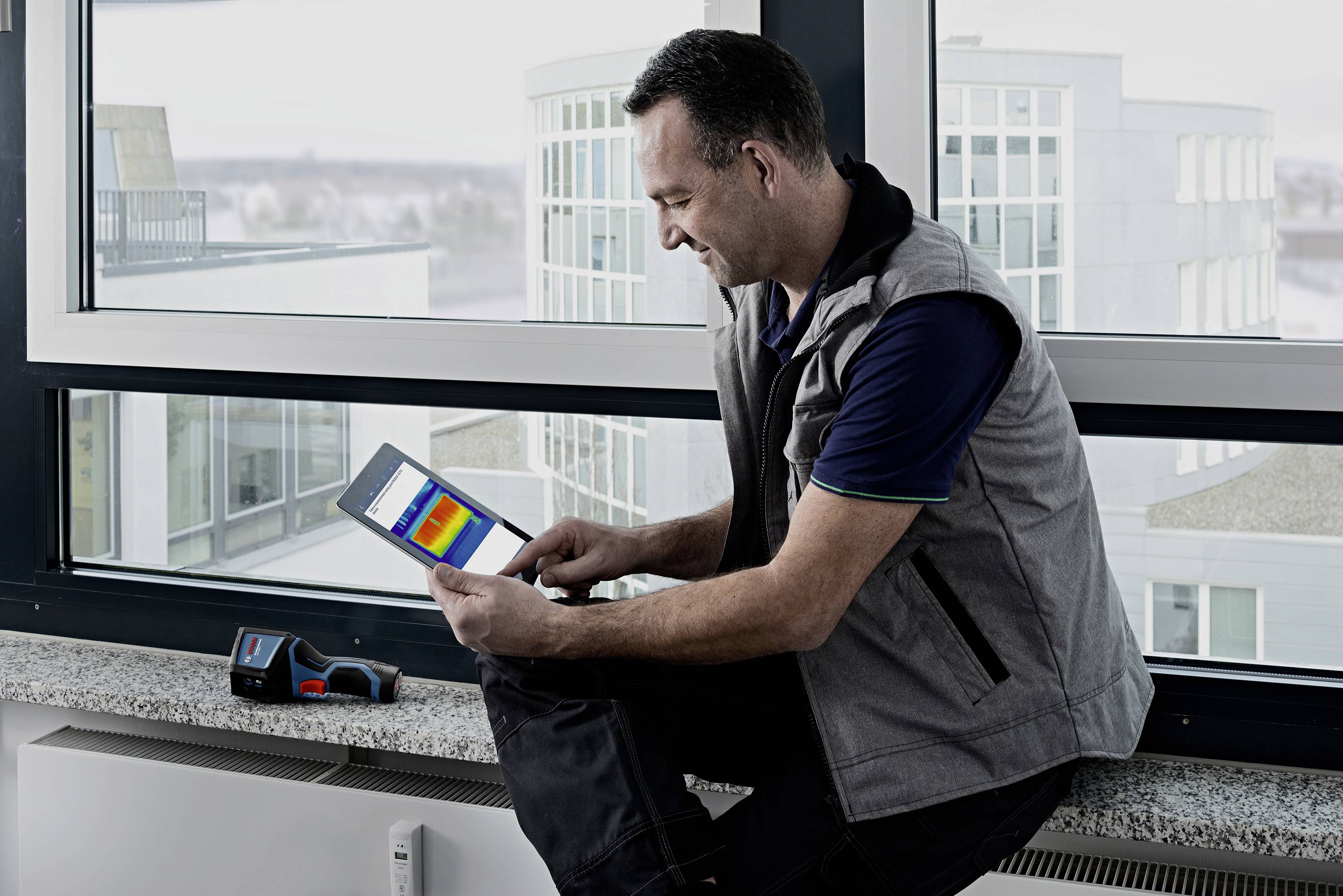 Un homme en tenue de travail est assis sur le rebord de la fenêtre et regarde un thermogramme sur une tablette, avec un appareil de mesure à côté.
