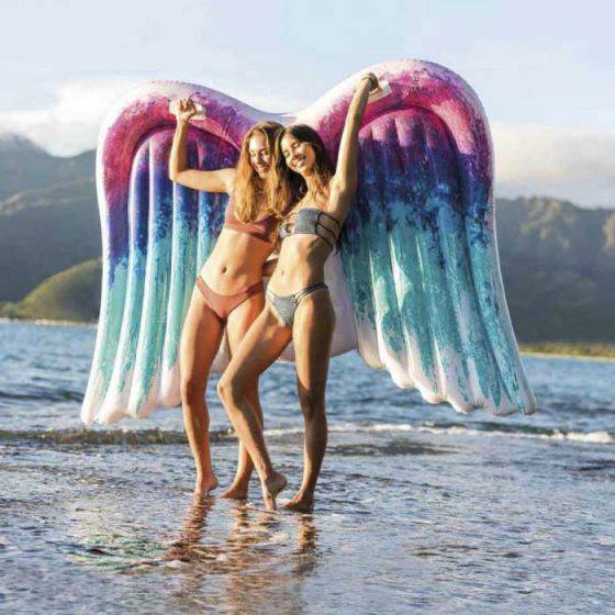 Deux personnes en tenue de bain posent sur une plage avec un grand accessoire ailé coloré derrière elles.
