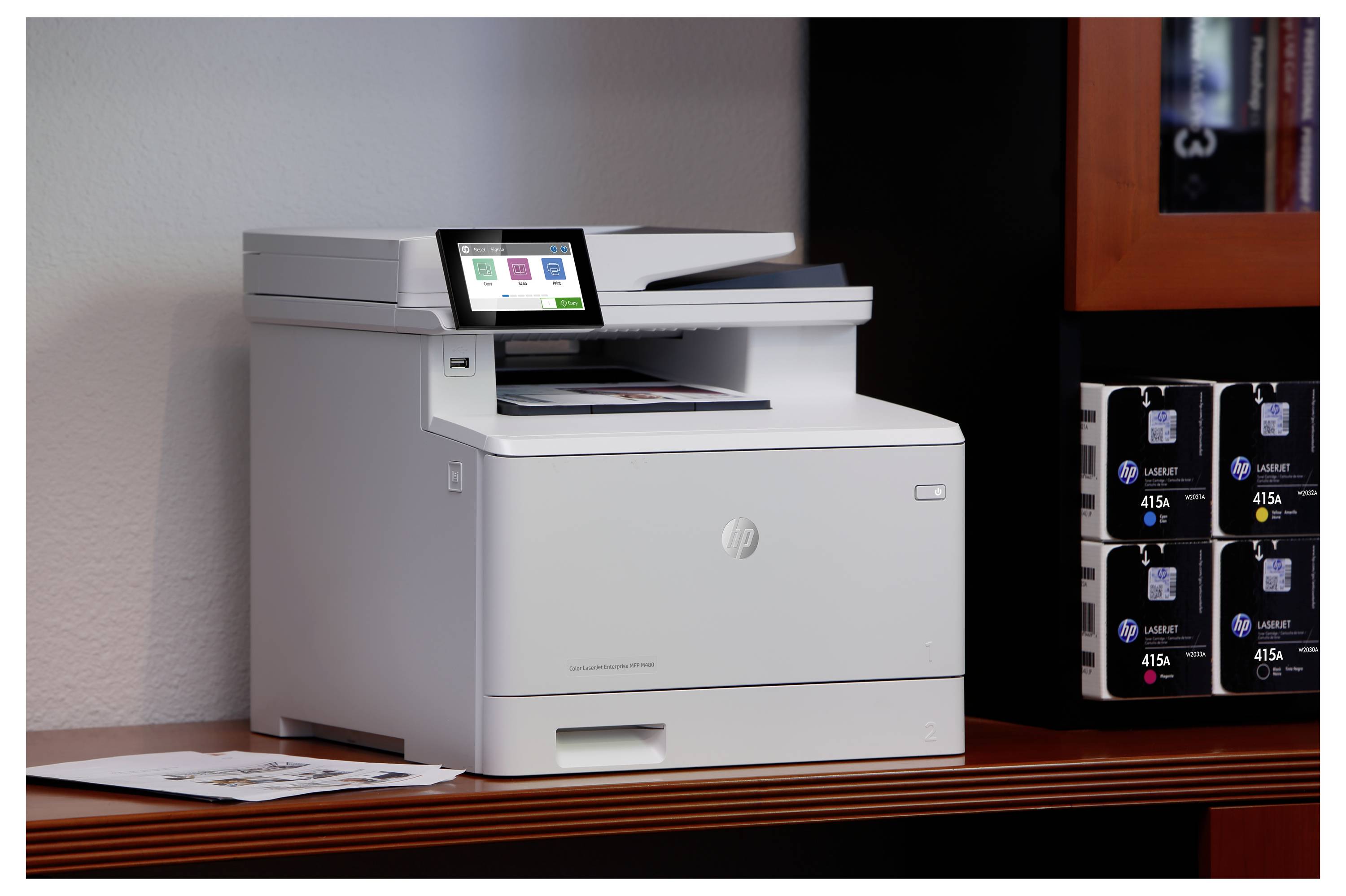 Imprimante multifonction HP blanche sur une étagère en bois, avec un panneau tactile visible et des boîtes de cartouches d'encre empilées à côté.