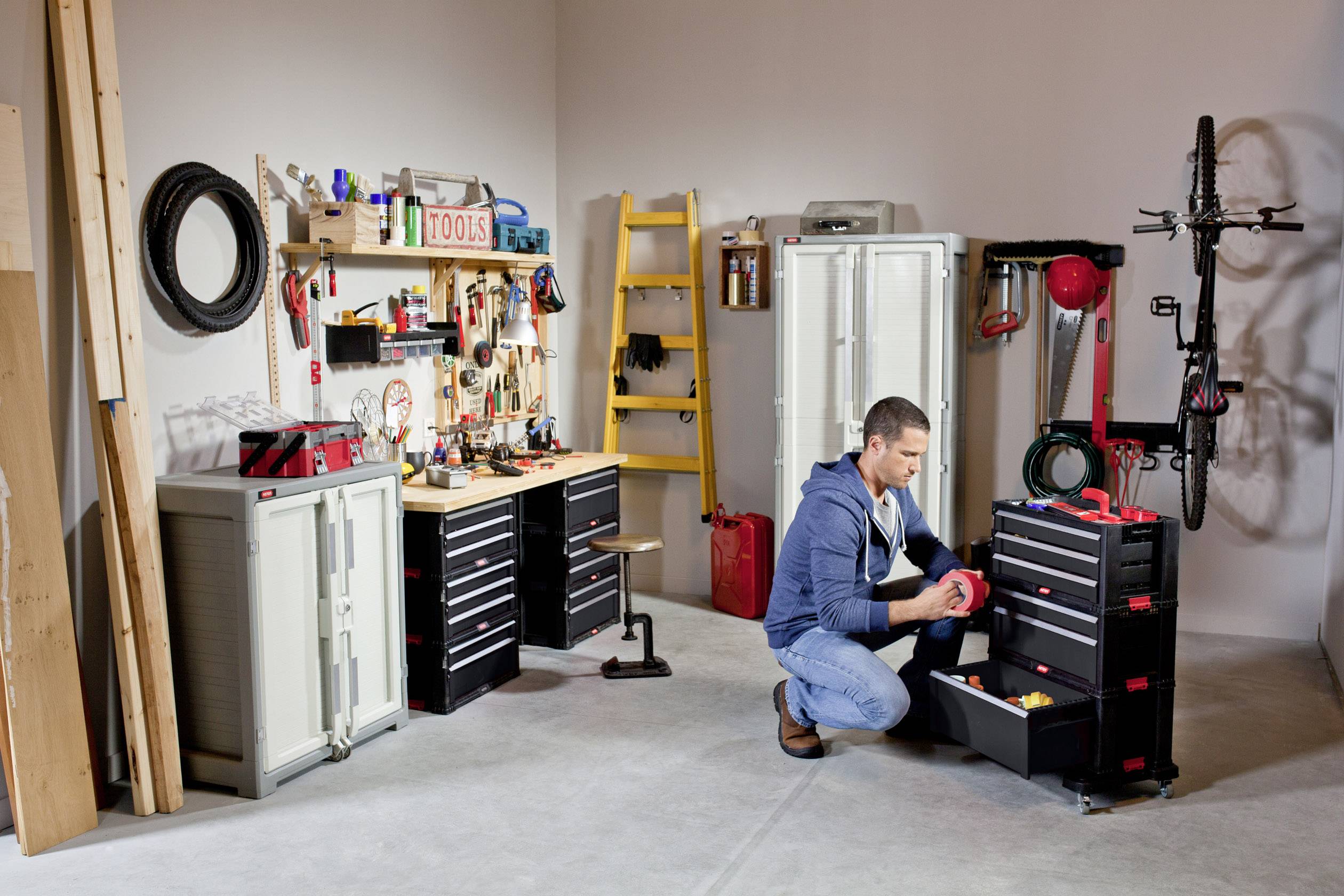 Un homme trie des outils dans un garage. Des étagères avec des outils et des options de rangement sont visibles en arrière-plan. Un vélo est accroché au mur.