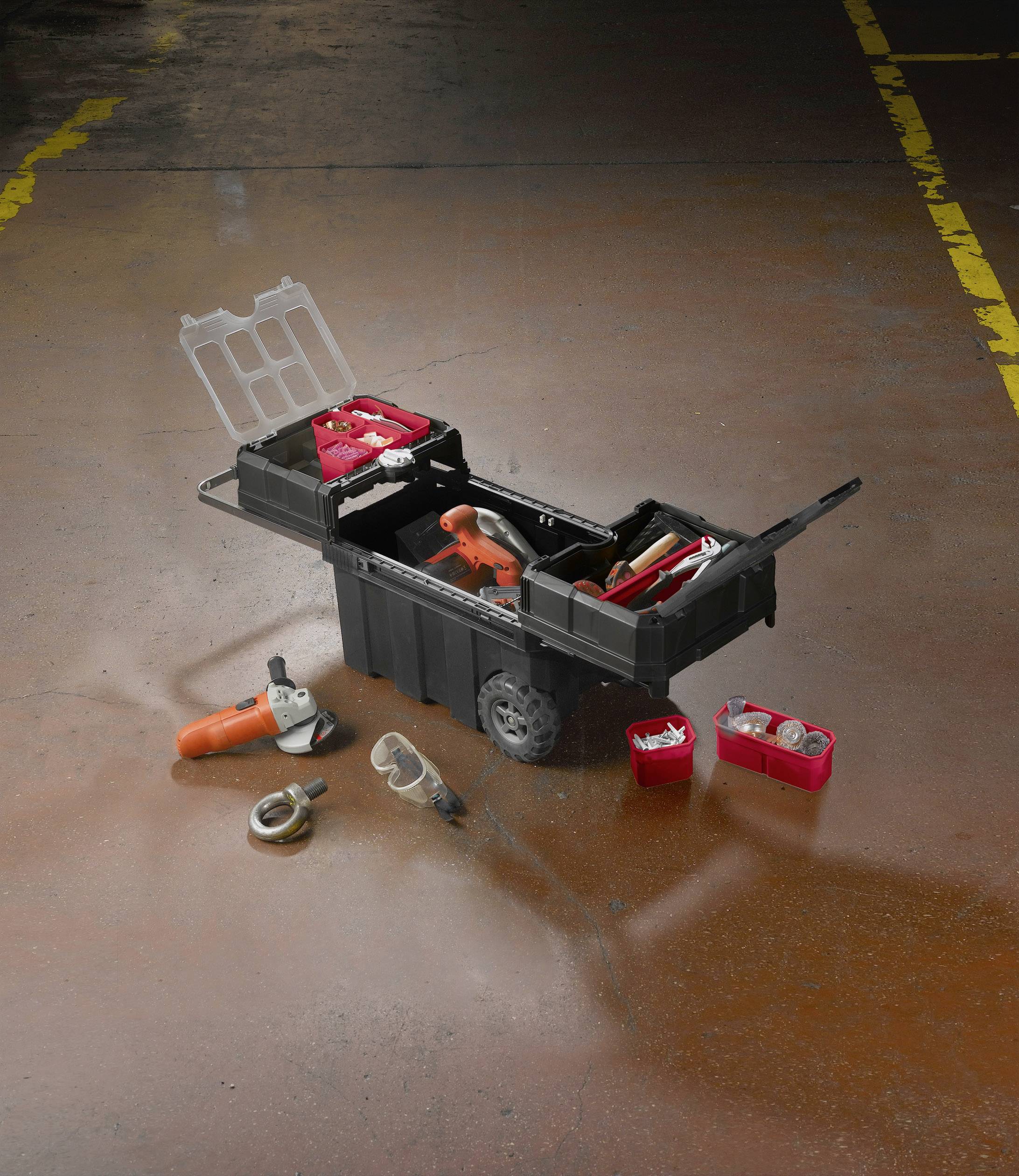 Une boîte à outils mobile ouverte avec divers outils, comprenant une meuleuse d'angle et des boîtes de petites pièces, posée sur un sol en béton.