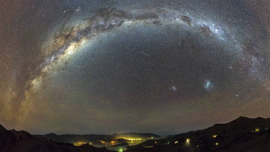 Un panorama du ciel nocturne montre la Voie lactée en arc lumineux au-dessus d'un paysage parsemé de lumières urbaines scintillantes.
