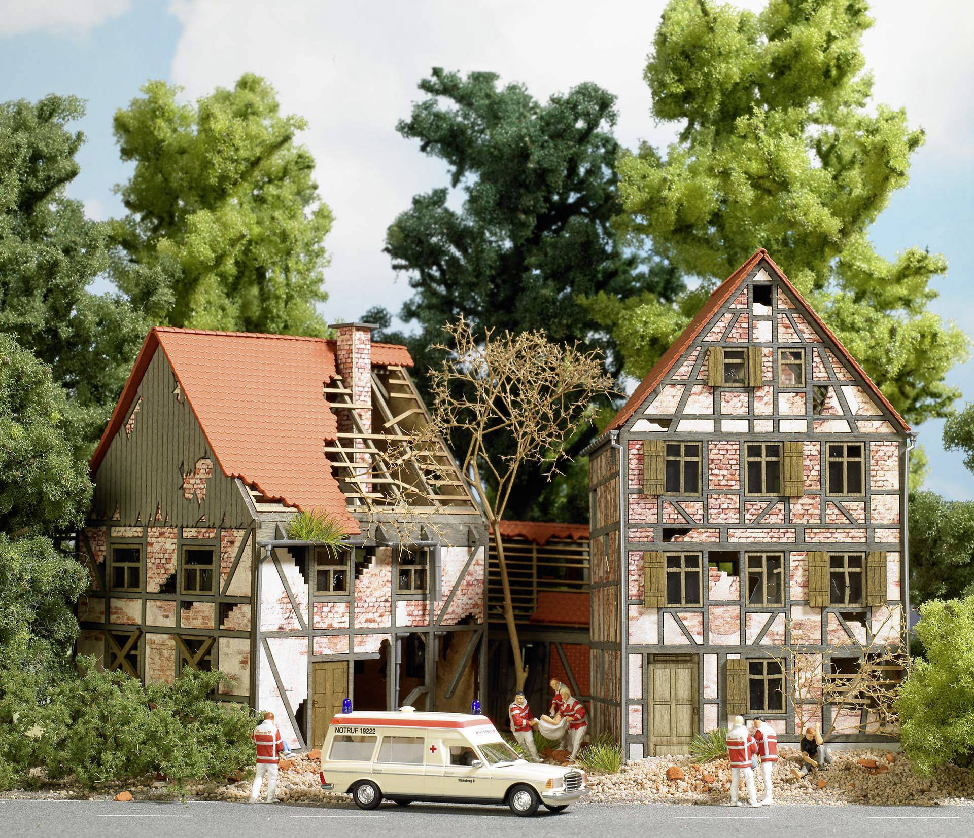 Une maquette d'une ancienne maison à colombages avec un toit endommagé. Devant se trouvent des figurines miniatures portant des uniformes rouges et une ambulance.