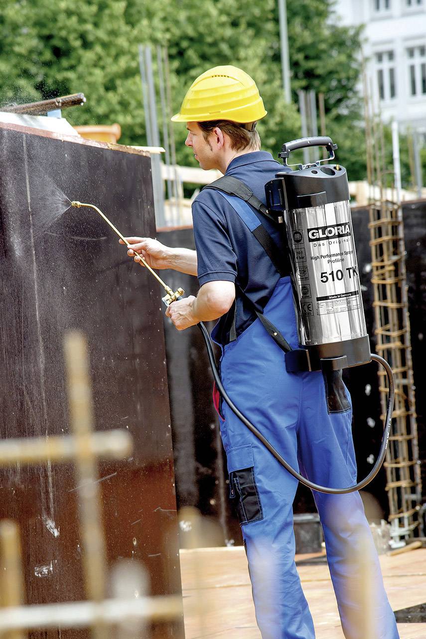 Un ouvrier en combinaison de protection et casque pulvérise un conteneur noir sur un chantier. Il porte un pulvérisateur dorsal portant l'inscription « GLORIA 510 T ».