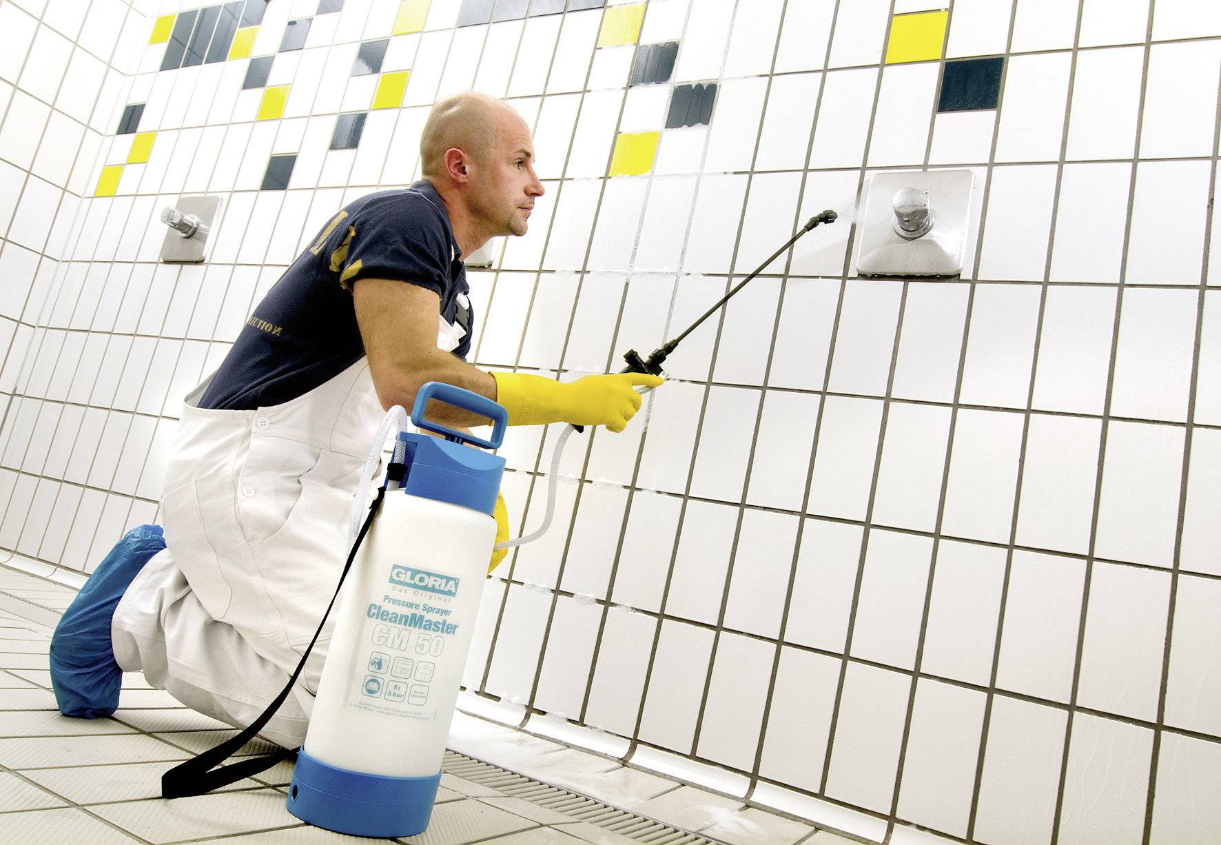 Un homme en tenue de travail nettoie des carreaux dans une salle de bain à l'aide d'un pulvérisateur.