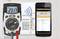Un multimètre numérique affiche 231,7 volts à côté d'un écran de smartphone avec des relevés de mesure. Un tableau électrique se trouve en arrière-plan.