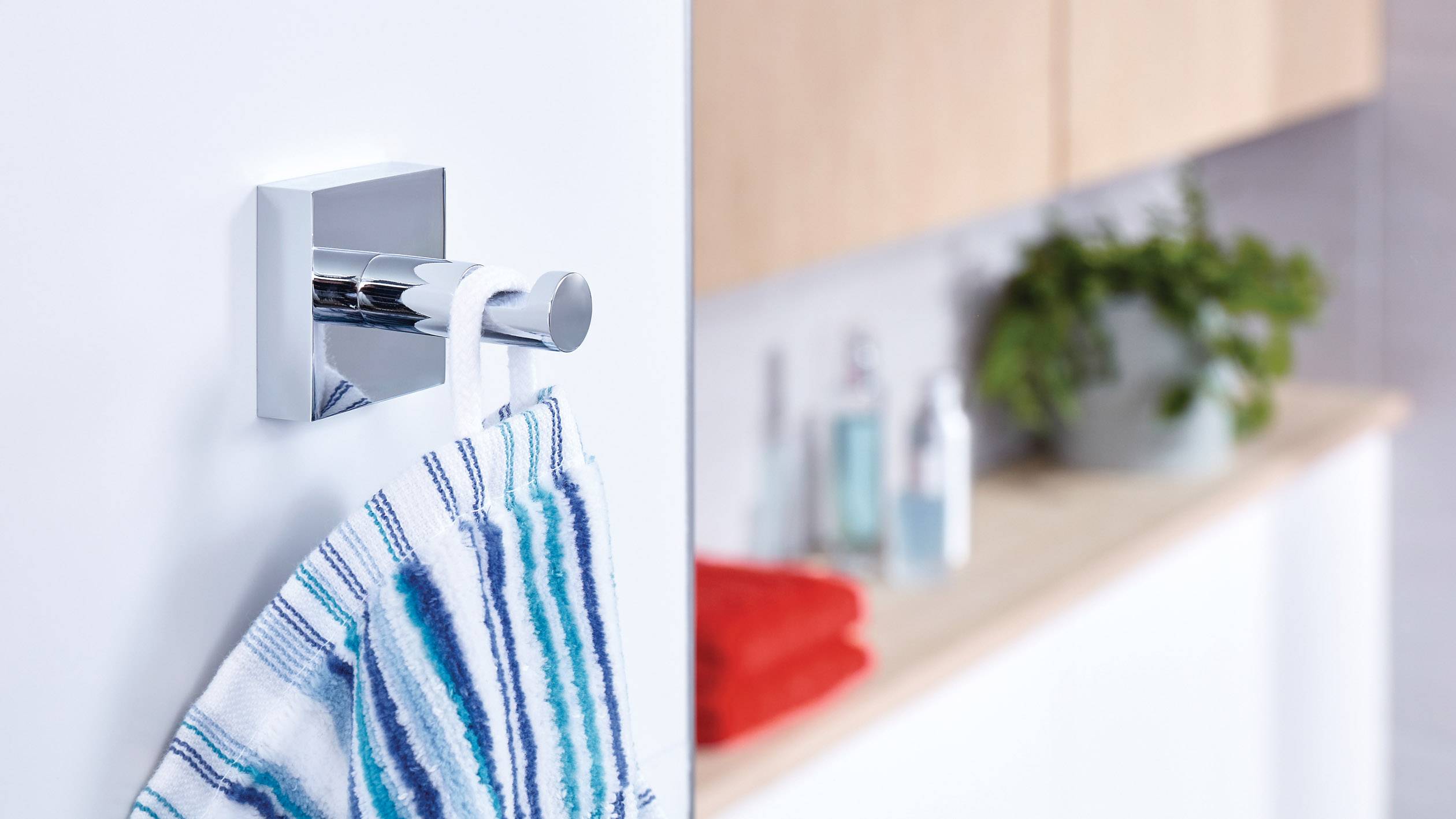 Une serviette est suspendue à un crochet mural moderne en argent dans la salle de bain. En arrière-plan, on distingue des produits de soin et une serviette rouge.