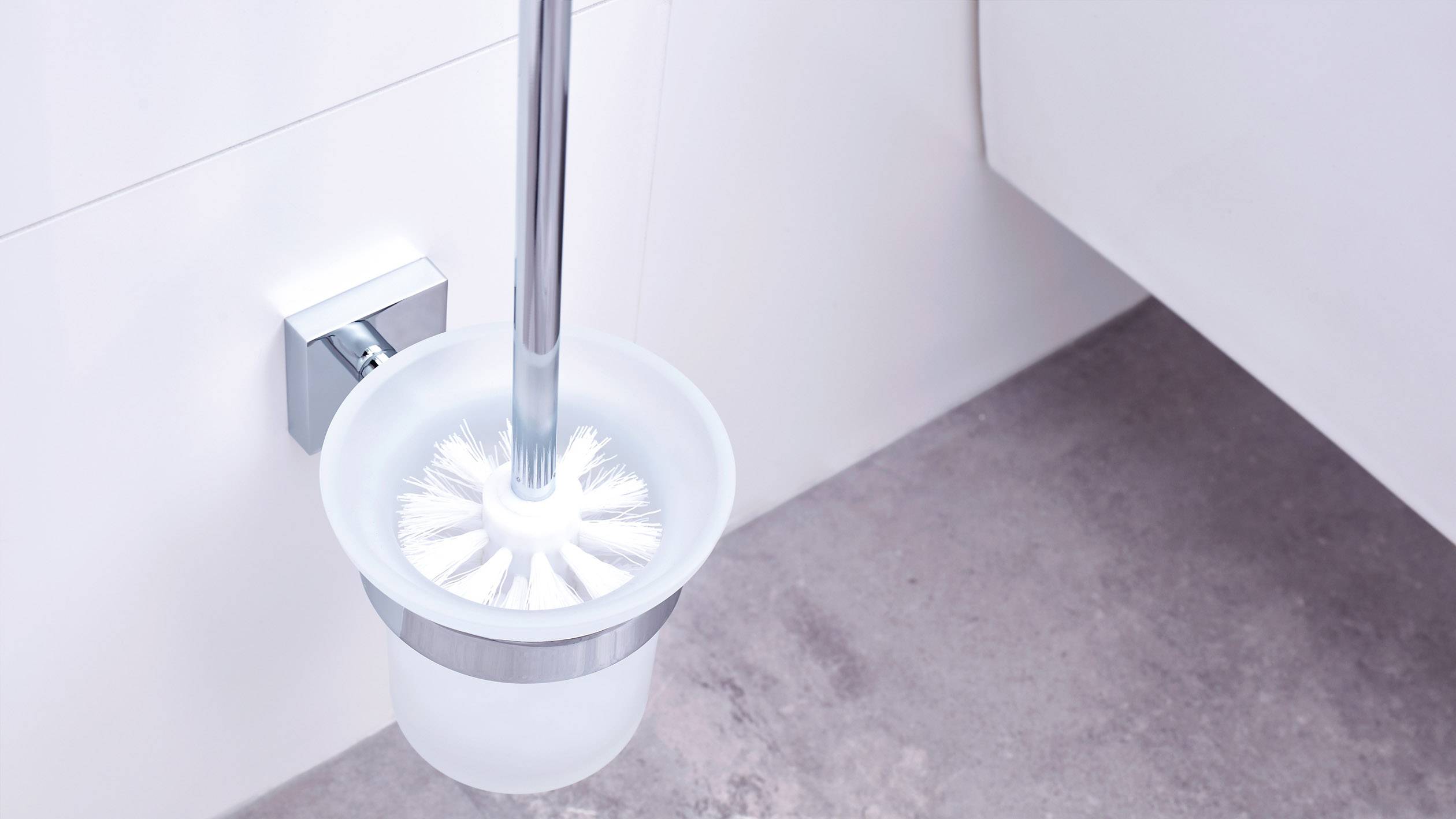 Une brosse de toilettes dans un récipient blanc est fixée au mur d'une salle de bains. Le sol est carrelé de gris.