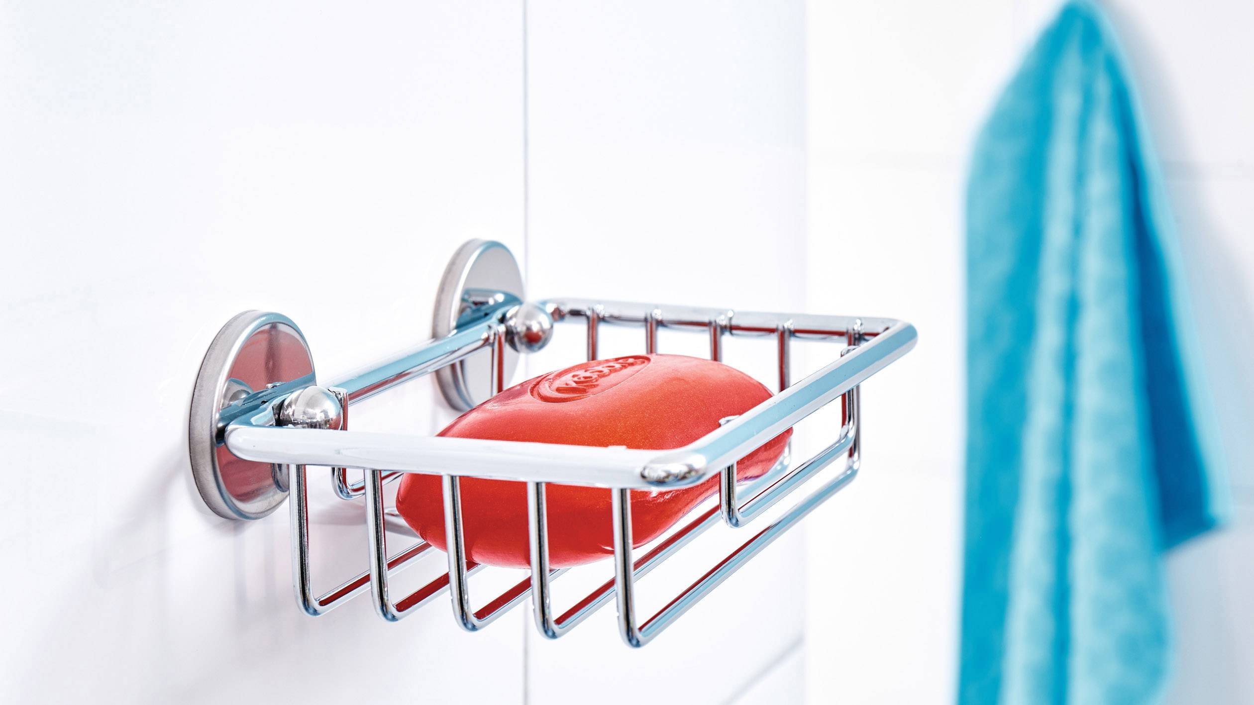 Support de savon en métal fixé au mur, avec un morceau de savon rouge. Un serviette bleue est suspendue en arrière-plan.