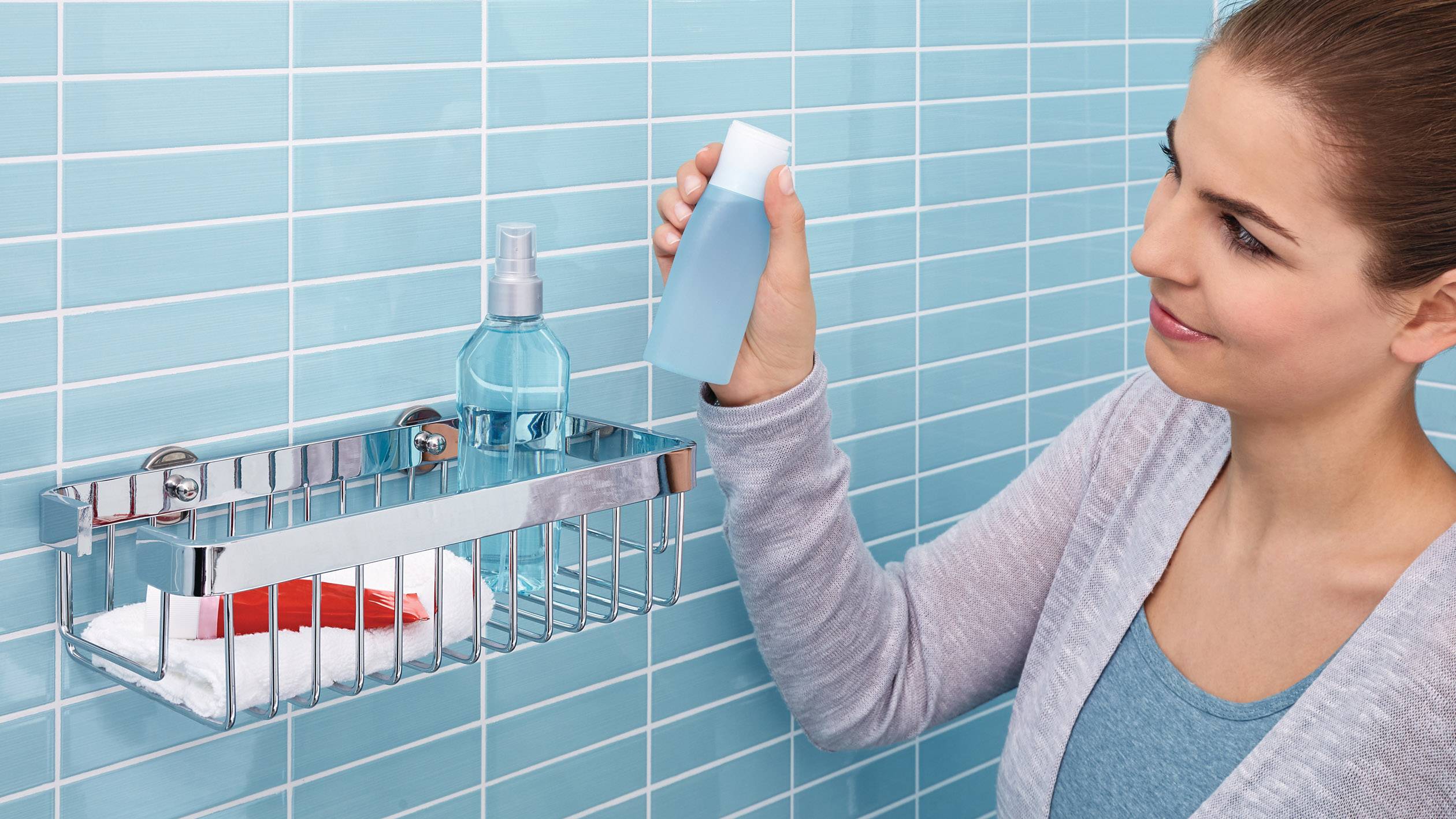 Une personne place une bouteille bleue dans une étagère métallique dans la salle de bain. Sur l'étagère se trouvent déjà un peigne rouge et un vaporisateur.