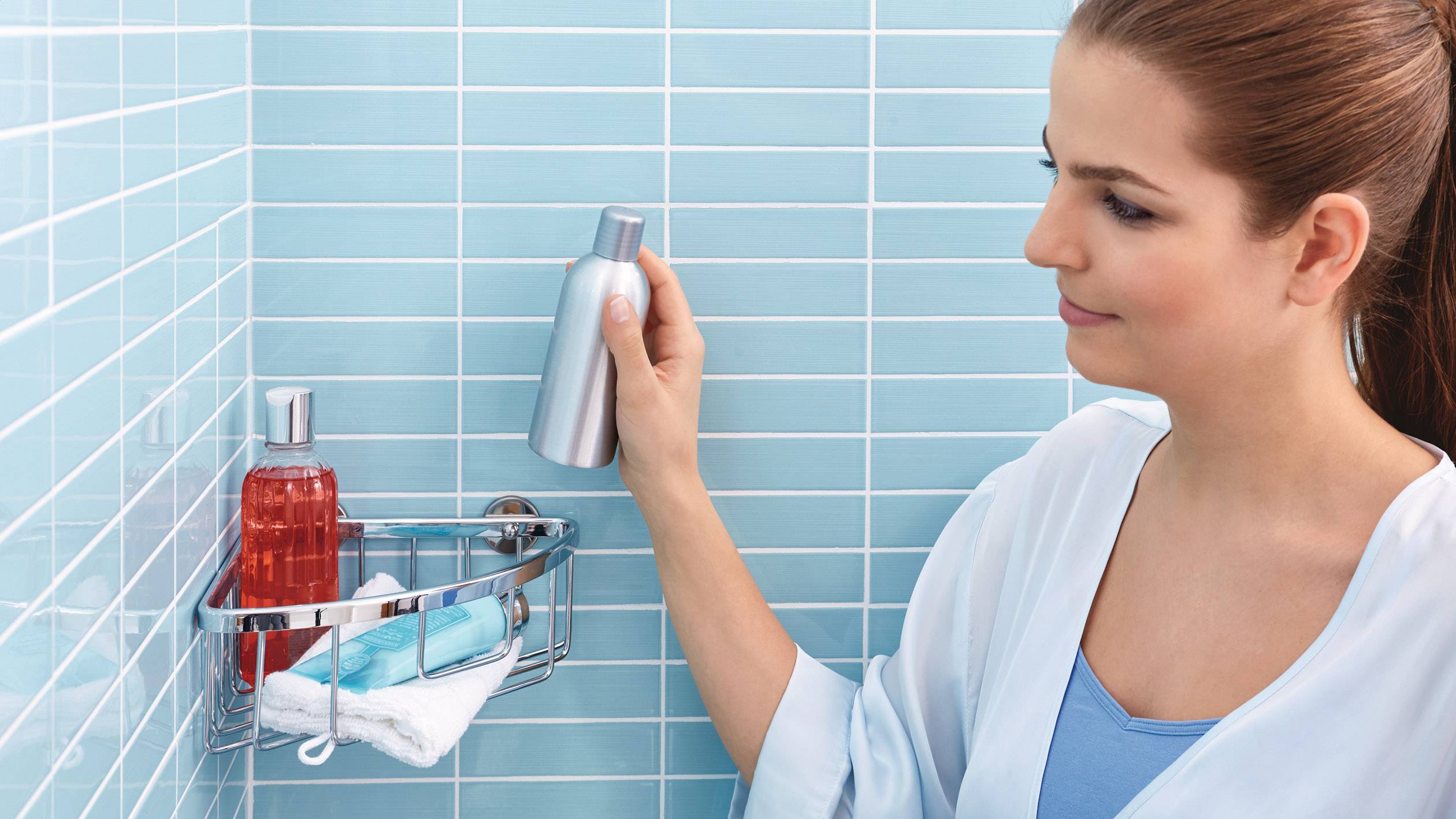 Une femme se tient dans une douche carrelée et saisit une bouteille de shampoing dans un panier de douche contenant divers produits de soin.