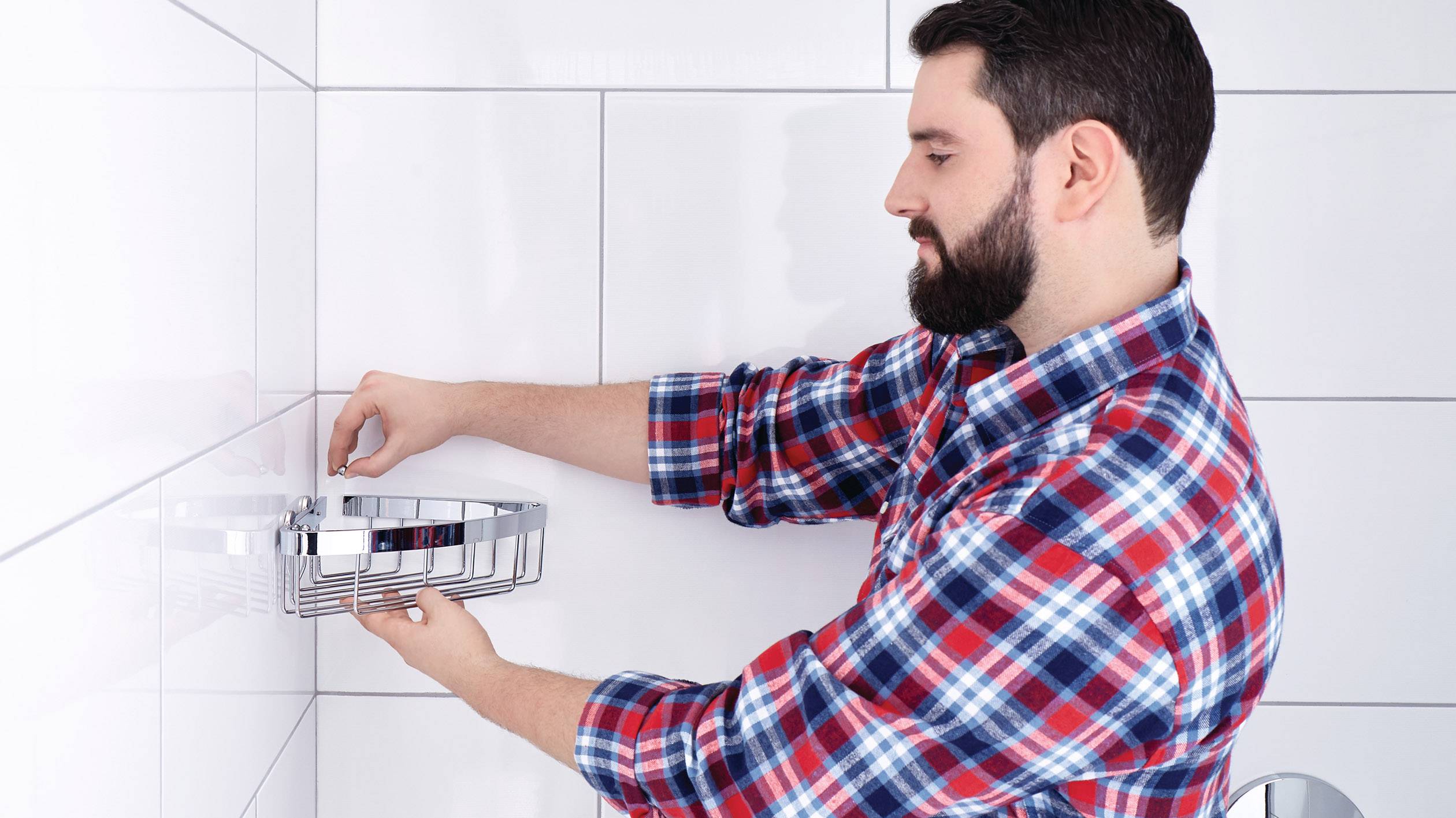 Un homme en chemise à carreaux installe une étagère de douche sur un mur de carrelage blanc.