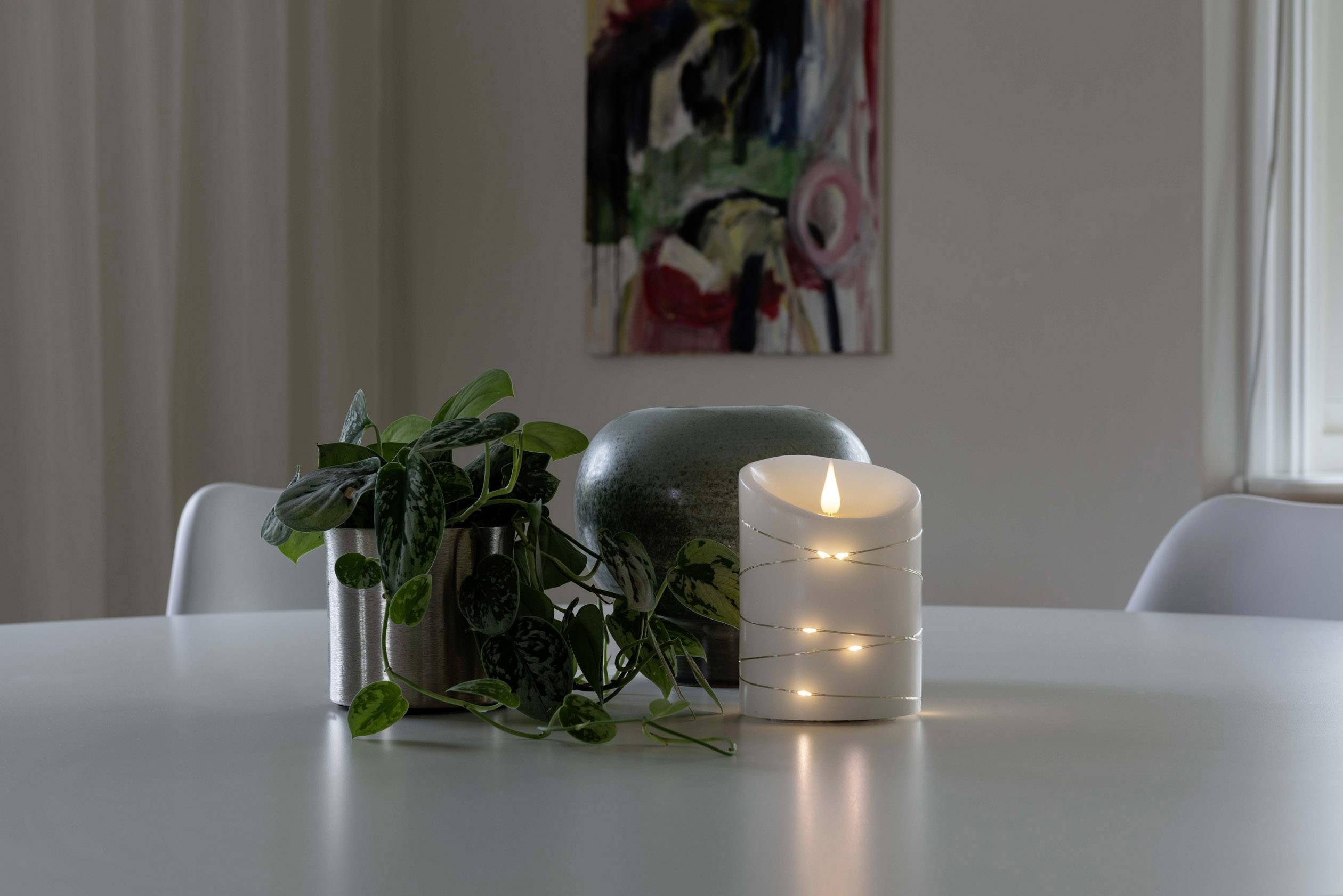Une table ronde avec une plante en trois pots et une bougie LED lumineuse. En arrière-plan, un tableau abstrait.