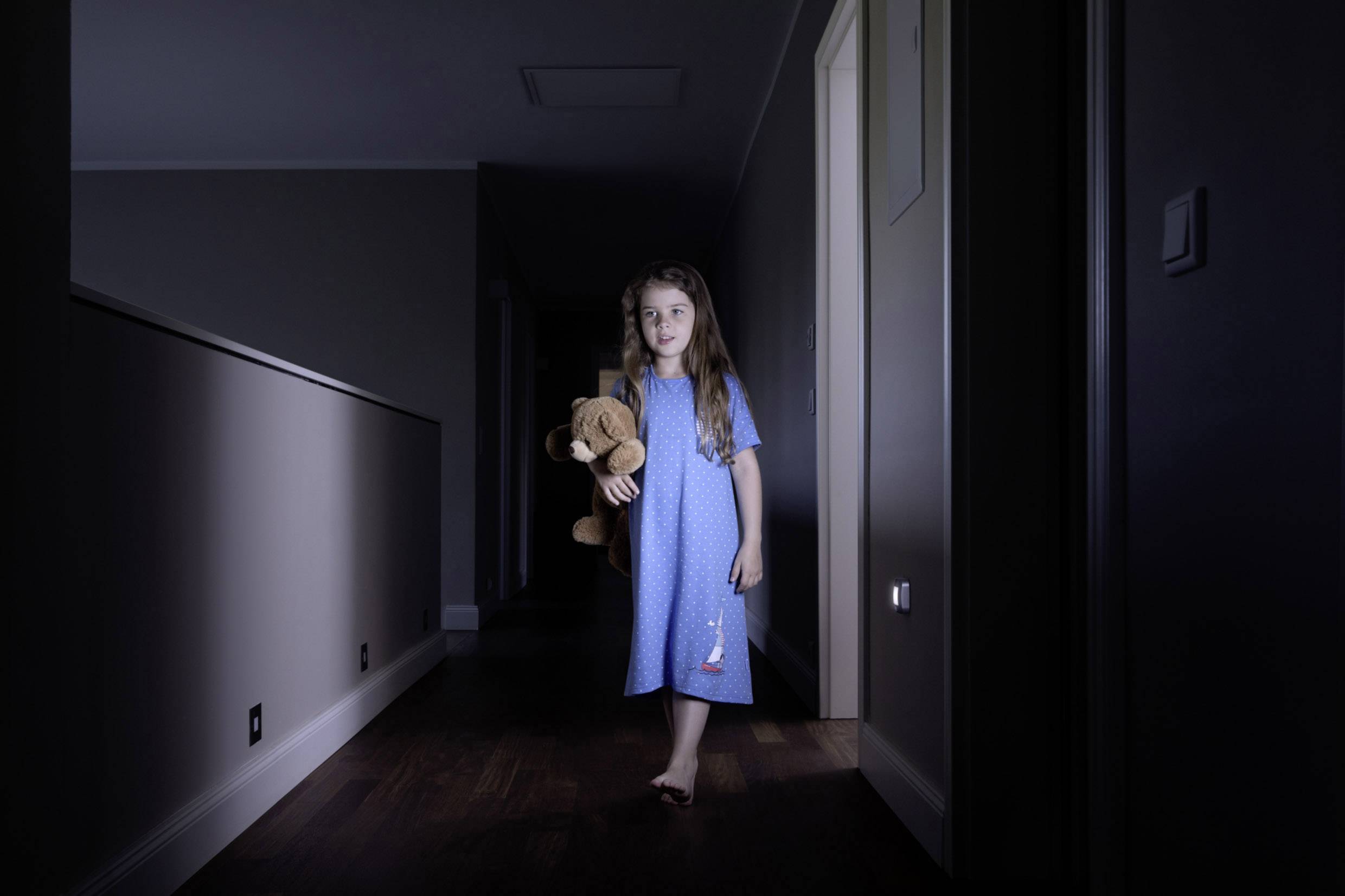Petite fille en chemise de nuit tenant un ours en peluche, marchant dans un couloir sombre.