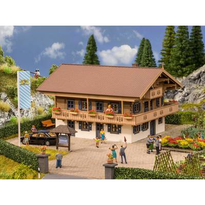 Une maison traditionnelle alpine avec un balcon orné de fleurs. Des personnes dans le jardin, des montagnes et des arbres en arrière-plan.