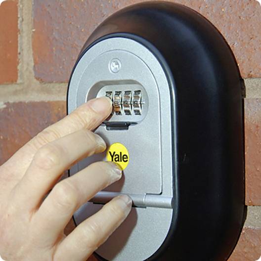 Une main entre une combinaison dans un coffre-fort à clés Yale avec des chiffres, installé sur un mur de briques.
