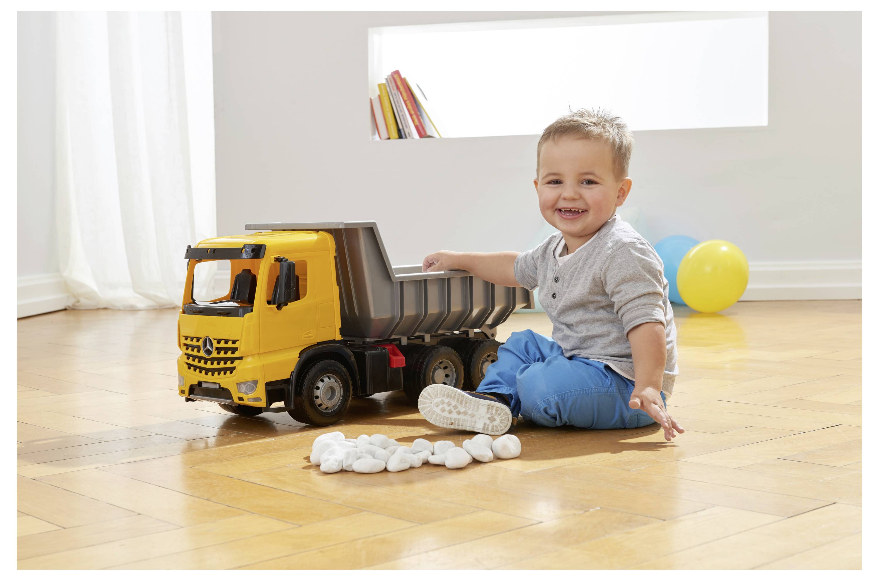Un jeune enfant souriant, assis sur un plancher en bois, jouant avec un camion-benne jaune et de petits galets blancs. Deux ballons se trouvent en arrière-plan.