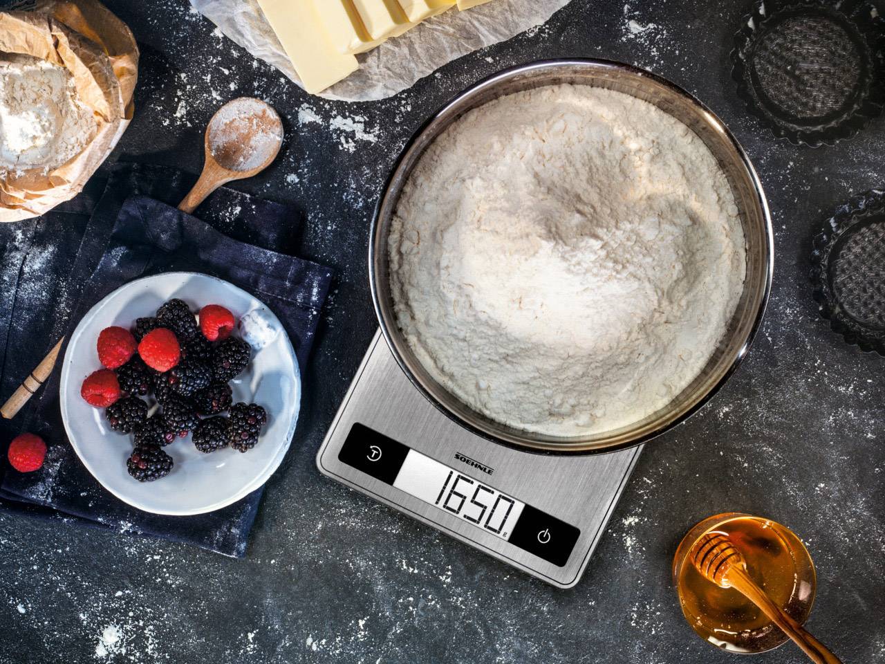 Un bol de farine sur une balance numérique affiche 1650 g, à côté des baies sur une assiette et un pot de miel sur une table.