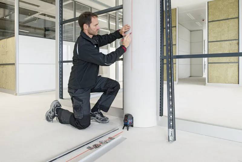 Un homme mesurant avec un niveau laser sur une colonne cylindrique dans un espace de bureau en cours de construction. Des outils et de l'équipement de mesure sont posés au sol.