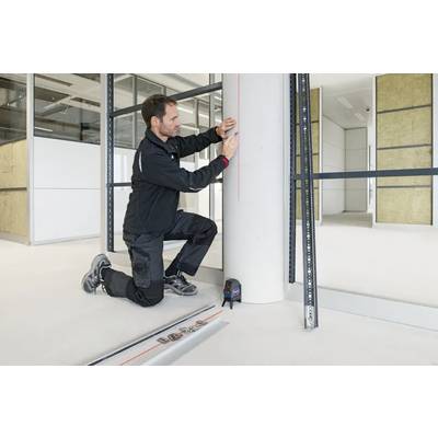 Un homme mesurant avec un niveau laser sur une colonne cylindrique dans un espace de bureau en cours de construction. Des outils et de l'équipement de mesure sont posés au sol.