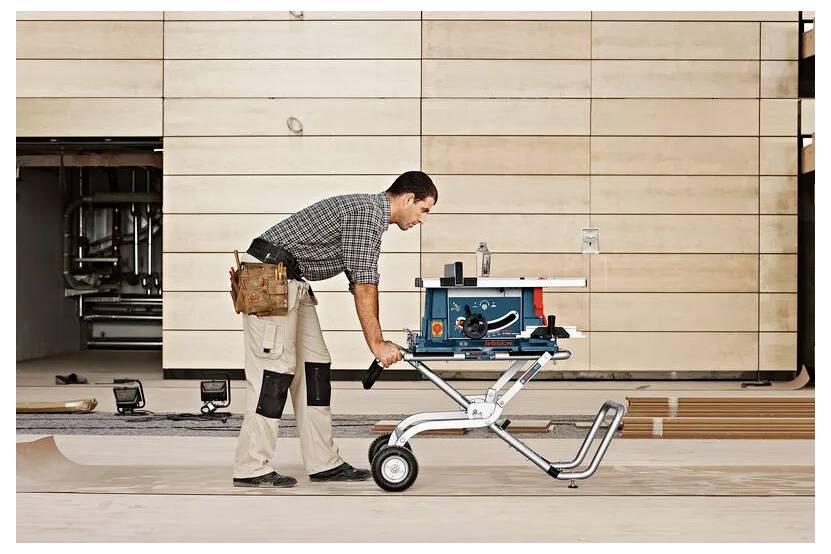 Un homme en tenue de travail pousse une scie à table portable bleue montée sur un support à roulettes à travers le sol d'un atelier, des outils visibles en arrière-plan.