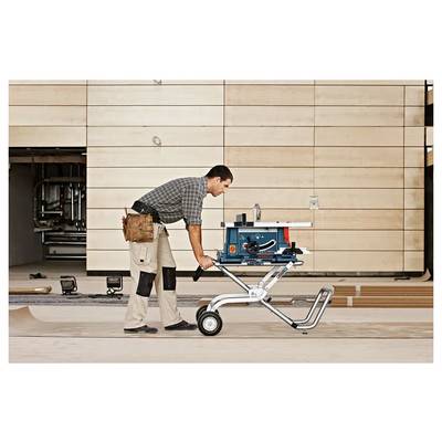 Un homme en tenue de travail pousse une scie à table portable bleue montée sur un support à roulettes à travers le sol d'un atelier, des outils visibles en arrière-plan.