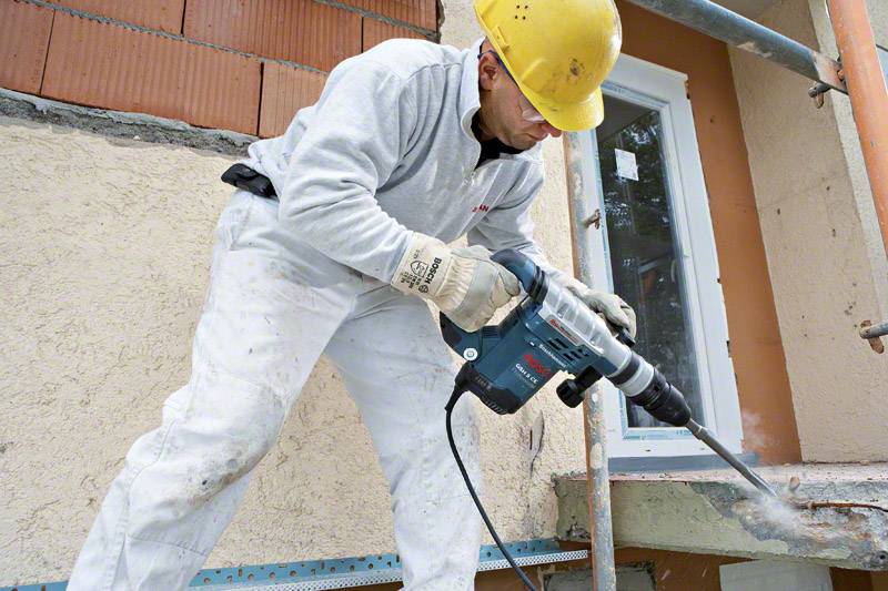 Un ouvrier en vêtements de protection et casque travaille avec un marteau-piqueur sur une surface en béton. Une fenêtre est visible en arrière-plan.