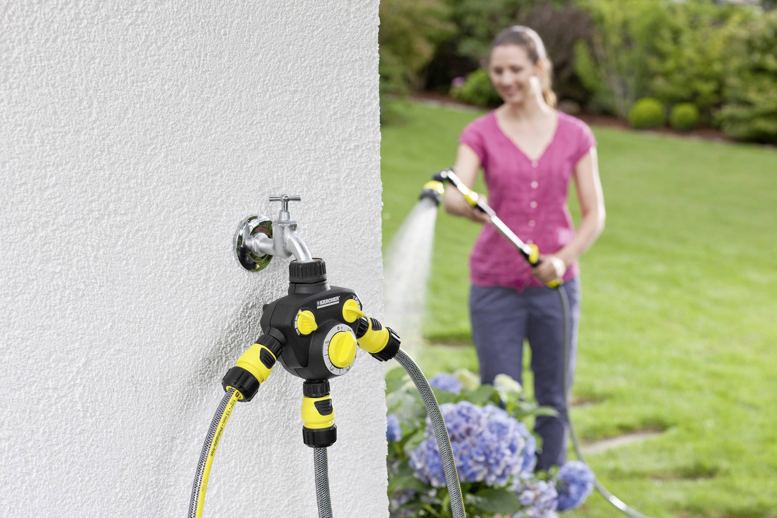 Une femme arrose le jardin avec un tuyau d'arrosage. Au premier plan, on peut voir un répartiteur d'eau fixé au mur avec plusieurs raccordements.