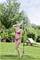 Une femme en bikini rose se tient sur une pelouse et prend une douche extérieure. Des arbres et un transat sont visibles en arrière-plan.