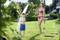 Un enfant en short de bain bleu se tient dans le jardin et se fait arroser par une femme en bikini avec un tuyau d'arrosage. Tous deux rient.